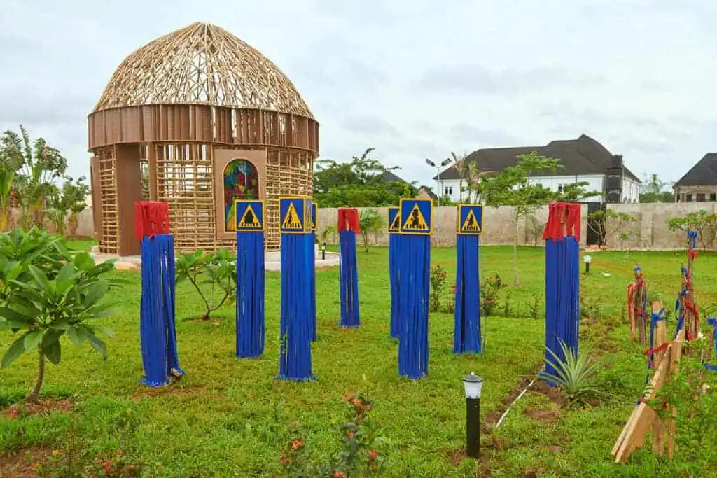 A garden with blue and red vertical poles, some topped with caution signs, stands in front of a dome-shaped wooden pavilion at The Black Muse Art Festival 2025. Houses and greenery are visible in the background under a cloudy sky.