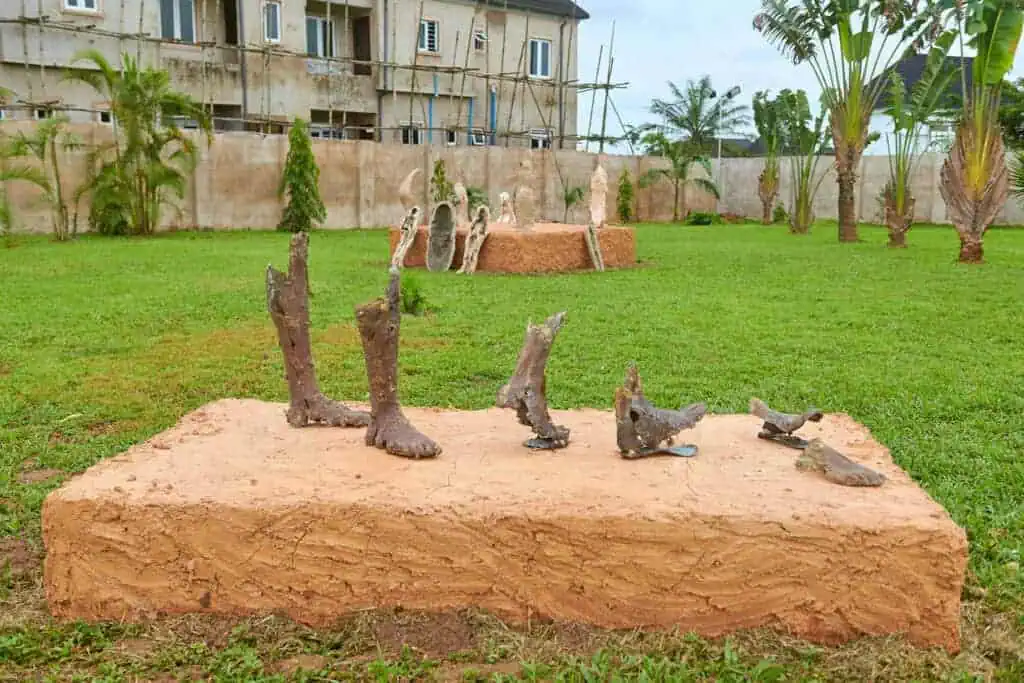 Sculptures of feet and lower legs are displayed on a clay pedestal in a grassy outdoor area at The Black Muse Art Festival 2025, with more sculptures and a partially constructed building in the background. Palm trees line the perimeter.
