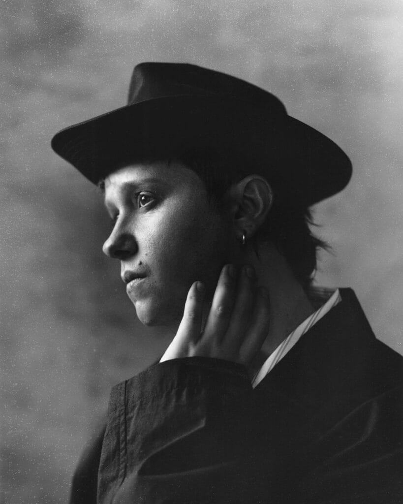 Black and white portrait of Conor Mason wearing a wide-brimmed hat and coat, gazing thoughtfully to the left. He touches his neck with one hand and wears a small hoop earring. The background is softly blurred.