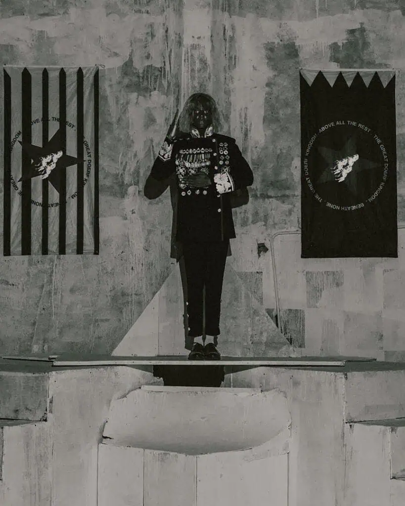 Stark black-and-white image of a figure positioned between graphic banners, evoking the symbolic and performative motifs linked to KABEAUSHÉ’s artistic narrative.
