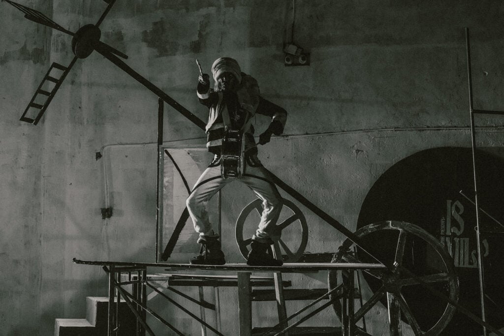 Figure posed defiantly on an industrial platform surrounded by mechanical wheels and scaffolding, captured in gritty monochrome lighting.