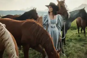 A horse girl in a light blue vintage-style dress and black hat stands in a grassy field among several horses, gazing into the distance with mountains visible in the background.