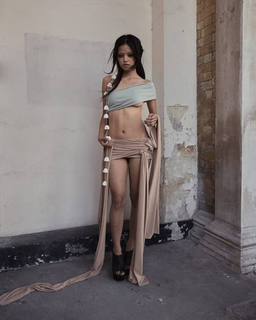 A woman stands in a worn, urban setting wearing asymmetrical beige and gray fabric by Jaoka Jaokha brand, draped around her body with white flowers strung along one side. She has a serious expression and wears black high-heeled shoes.