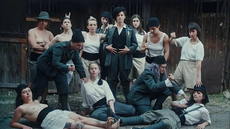 A group of people in vintage military-style and peasant clothing pose dramatically outdoors, evoking the spirit of 'Fiume o Morte!' by Director Igor Bezinović; some are shirtless, others brandish knives before a rustic wooden background.