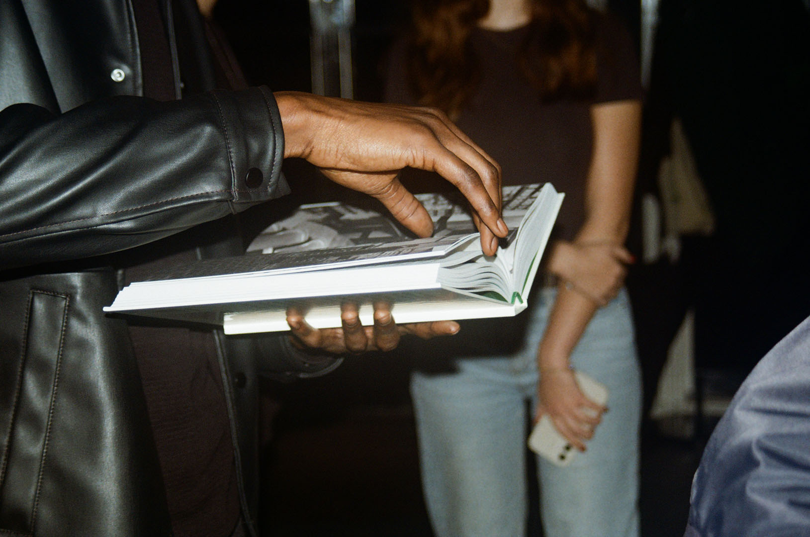 A person wearing a black jacket flips through a book, while another in a brown shirt, jeans, and classic CLARKS stands in the background holding a phone.
