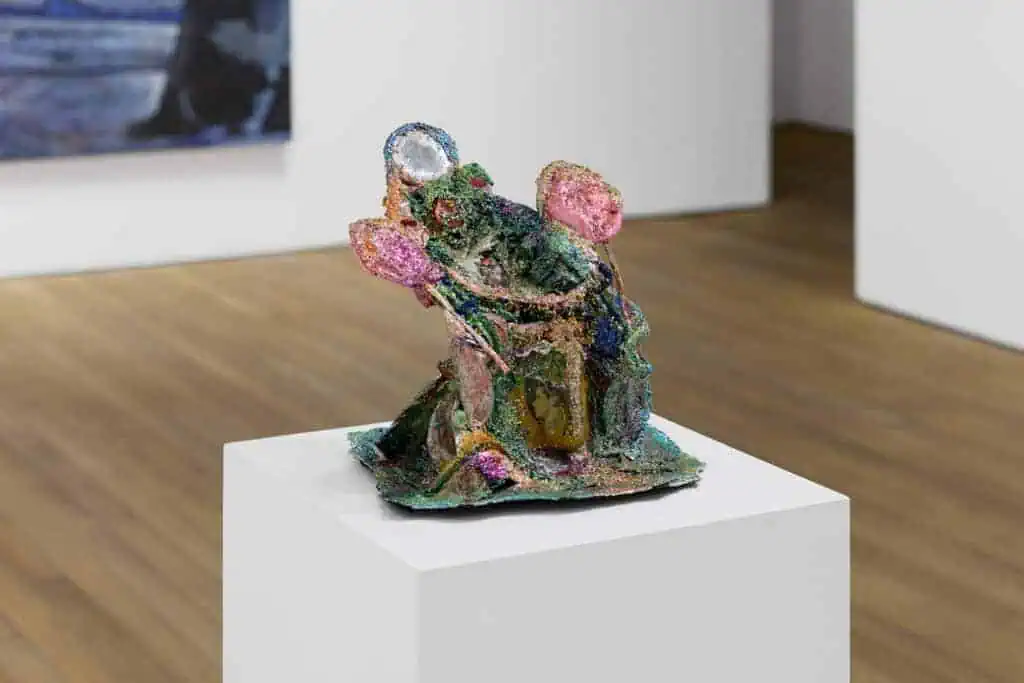 A colorful, abstract ceramic sculpture with glittery textures and iridescent sections sits on a white pedestal in Ginny on Frederick’s Frieze 2025 presentation, part of Crude Hints Towards curated by Charlotte Edey.