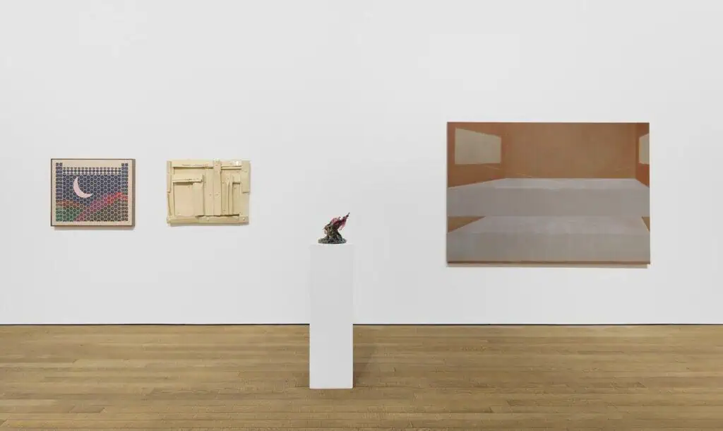 Three artworks hang on a white gallery wall at Crude Hints Towards, curated by Charlotte Edey at Ginny on Frederick for Frieze 2025. A grid moon piece, cream abstract relief, and minimalist bed painting are joined by a small dark sculpture on a white pedestal.