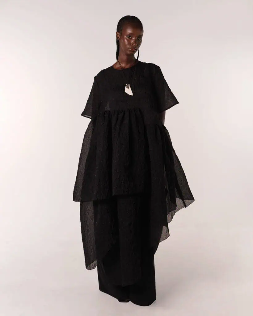 A person stands against a plain background wearing a layered, textured black dress designed by Moma Hilo. They have dark skin and a confident, calm expression.