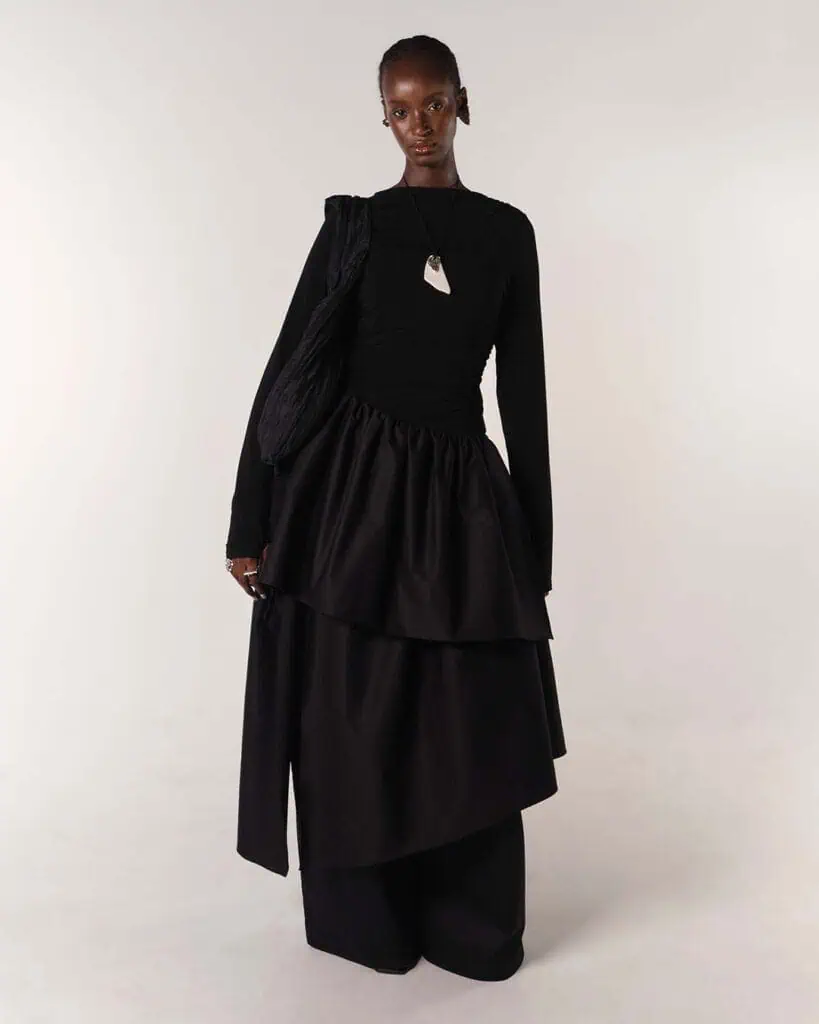 A person stands against a plain background wearing a long black layered skirt and fitted top—clothes designed by Moma Hilo—a large sculptural bag, and a bold white geometric necklace. They have a neutral expression and minimalist styling.