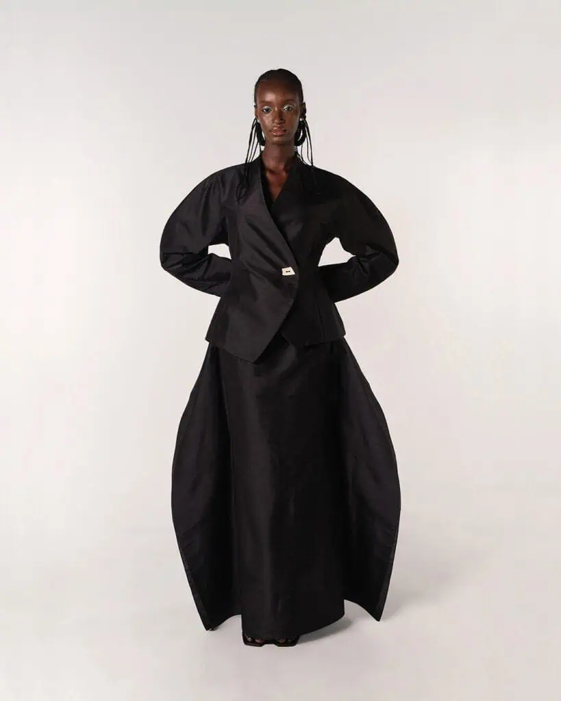 A woman stands against a plain background wearing striking clothes designed by Moma Hilo—a black suit with exaggerated, sculptural sleeves and a long, structured skirt that flares at the sides. She looks confidently at the camera with hands on her hips.