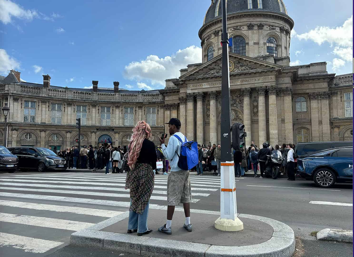 Two people stand at a crosswalk facing a large historic building with a dome, reminiscent of the grandeur seen at the Vivienne Westwood SS26 Runway. A crowd gathers near the entrance under a partly cloudy sky, with cars parked along the street.