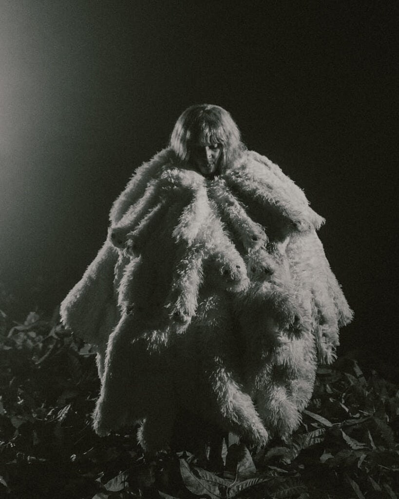 Figure wrapped in a voluminous white fur-like garment, standing outdoors in low light with a theatrical, surreal atmosphere.