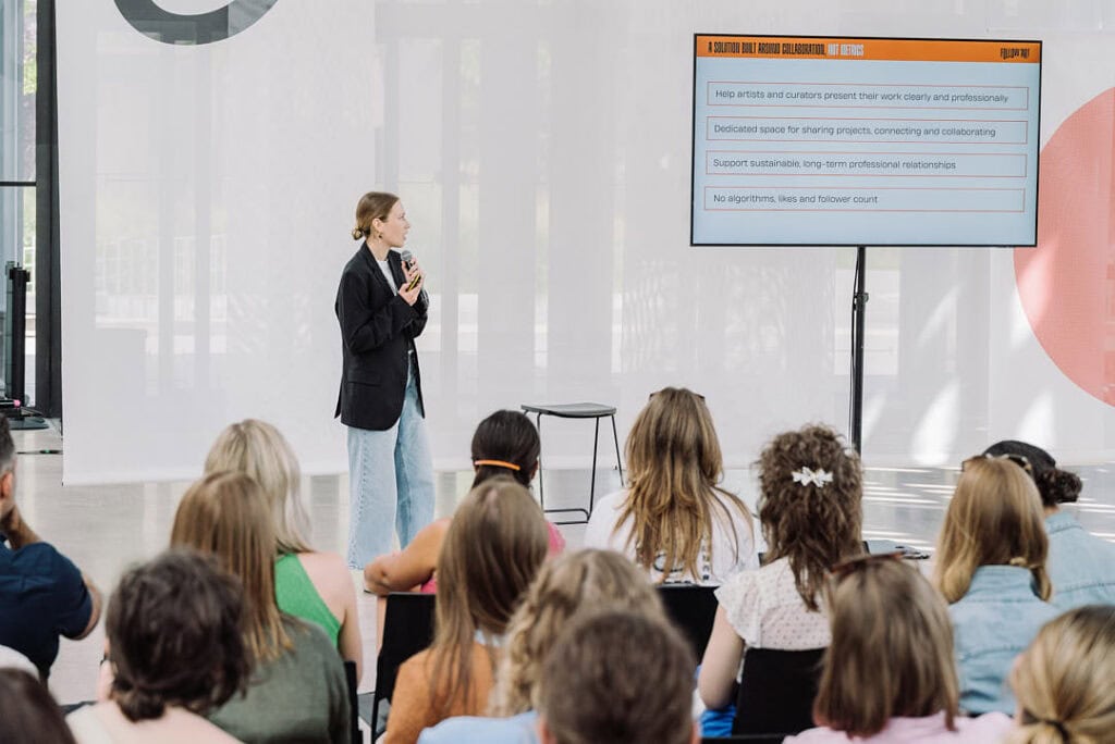 A speaker stands on stage with a microphone, presenting to an audience. A large screen displays FOLLOW.ART: A New Professional Tool for Artists and Curators, listing its principles. The room has bright natural light and modern decor.