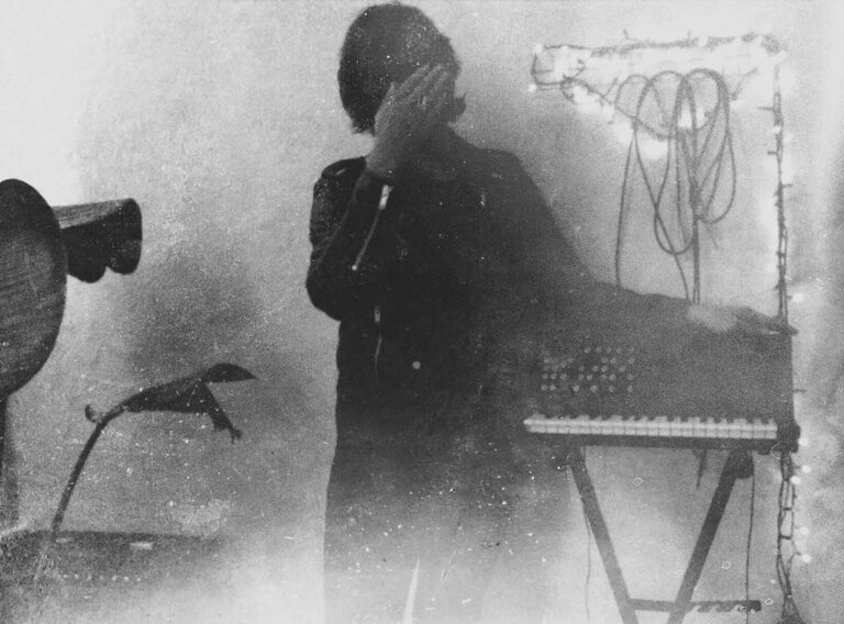 Public Memory performing on a vintage synthesizer in a dim, grainy room, evoking trip-hop and ambient tones.