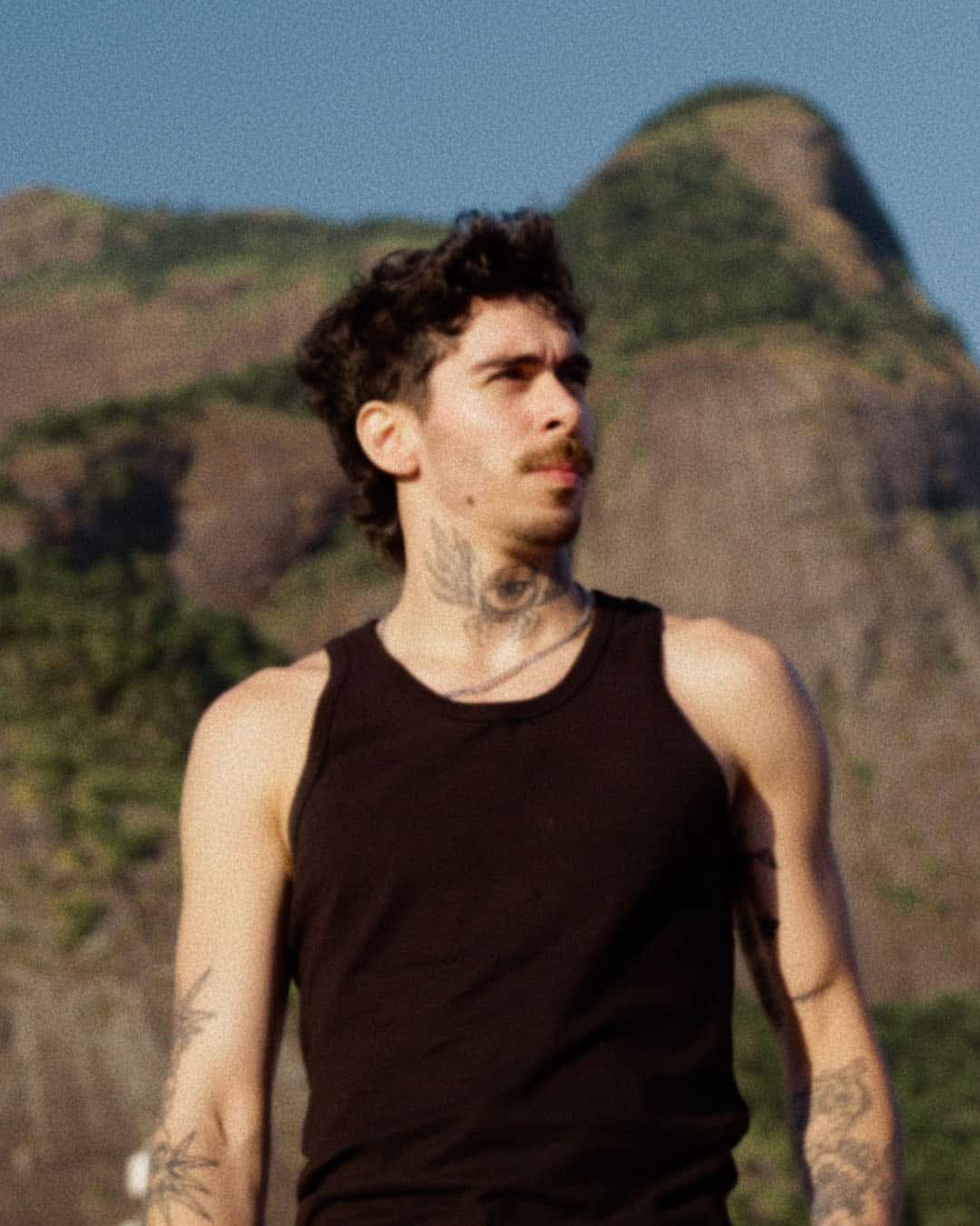 Mochakk, wearing a black sleeveless shirt, stands outdoors with mountains in the background. The natural lighting and clear sky evoke a scene reminiscent of a still from Belo horizonte documentary.