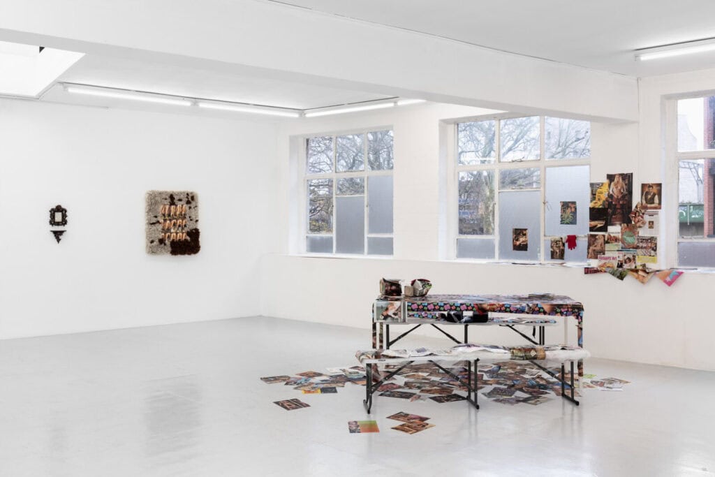 A bright, white-walled gallery room at Rose Easton Gallery displays a patterned table and bench covered with art supplies and scattered magazines. Artworks and photos—part of ‘Abigail’s Party’—adorn the walls, windows, and floor.
