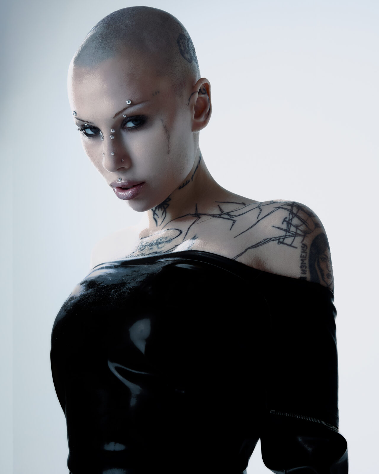 Siiickbrain for The cold magazine printed issue with a shaved head, facial piercings, and visible tattoos on shoulders and chest, poses in a shiny black off-the-shoulder outfit. An intense expression is lit dramatically from the side.