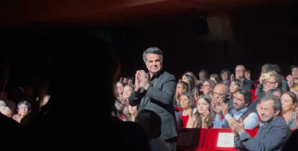 Jafar Panahi in a black suit stands and claps in the middle of a crowded, dimly lit theater audience. Others around him are seated and also applauding, spotlights highlighting the moment.