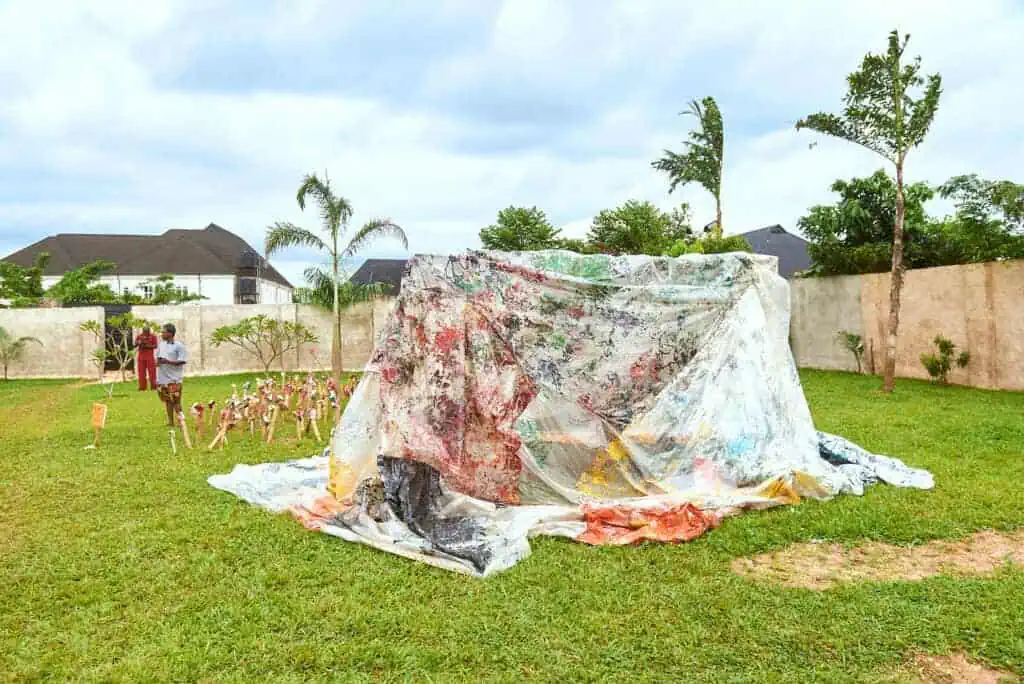 A large, colorful, painted sheet draped like a tent sits on grass in a yard at The Black Muse Art Festival 2025, surrounded by a few trees and a concrete wall. Two people stand near a small display of objects to the left, with houses visible in the background.