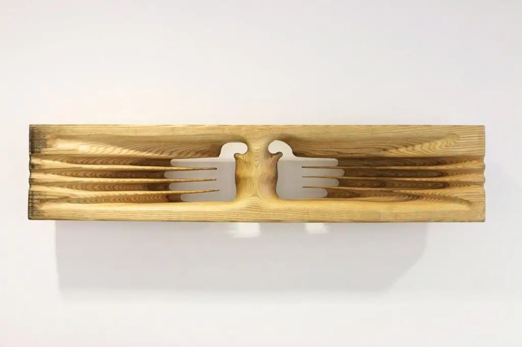 A wooden plank with two stylized bird shapes carved out, facing each other. Inspired by The Hari Art Prize’s celebration of London’s emerging artists, the wings form parallel grooves that extend outward along the wood grain in a symmetrical pattern.