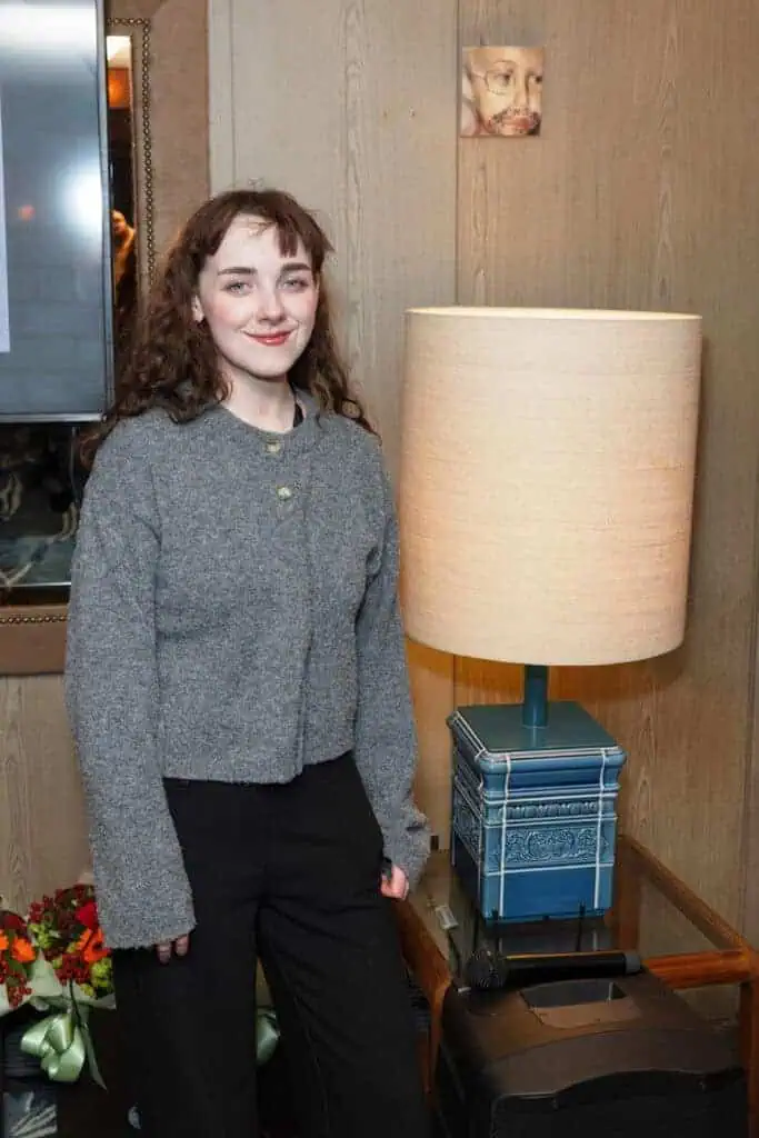 A young woman with wavy brown hair, wearing a gray sweater and black pants, stands indoors next to a table lamp. In the background—framed art and flowers suggest an ambiance where The Hari Art Prize is celebrating London’s emerging artists.
