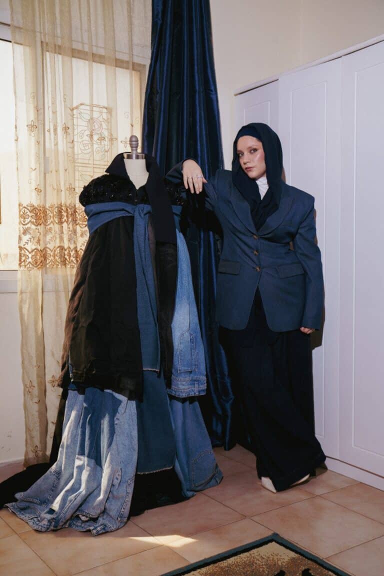 A person wears a blue hijab, oversized blazer, and wide-leg pants, standing beside a mannequin styled in jackets and denim skirts. The chic indoor setting hints at Samah Sulyman designer flair with cream walls, a curtain, and white wardrobe.