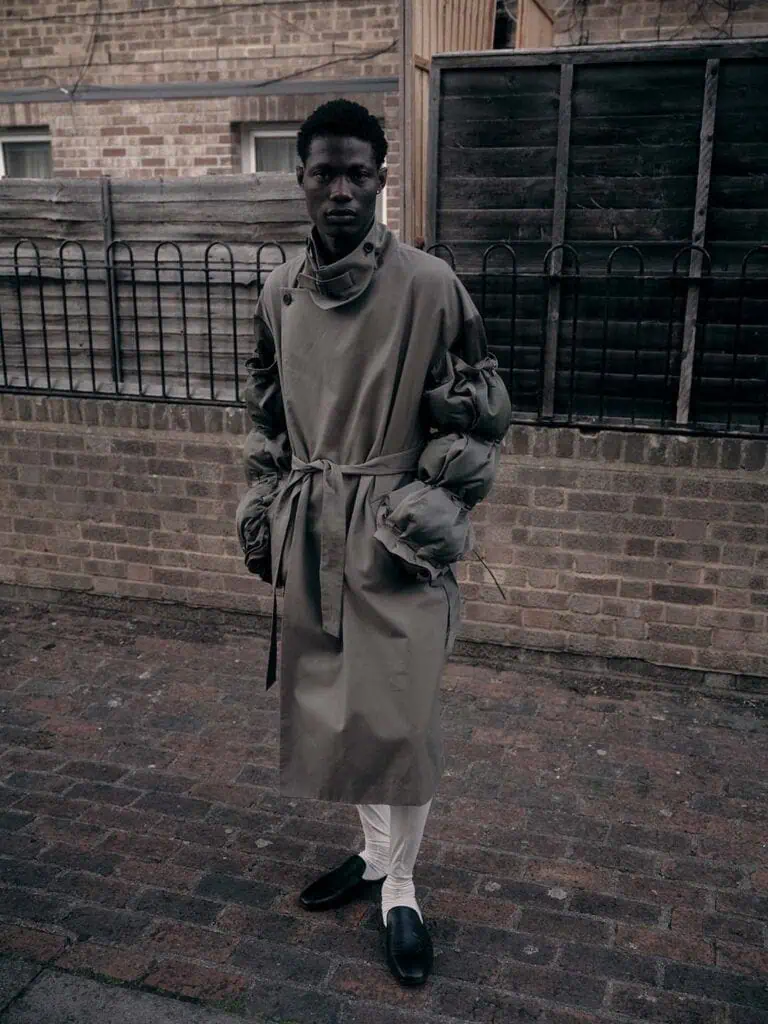 A person stands outside on a brick pavement wearing a long, belted, olive-green coat with puffy sleeves—clothes designed by Yonzu—white pants, and black shoes. The background features a brick wall, wooden fence, and metal railings.