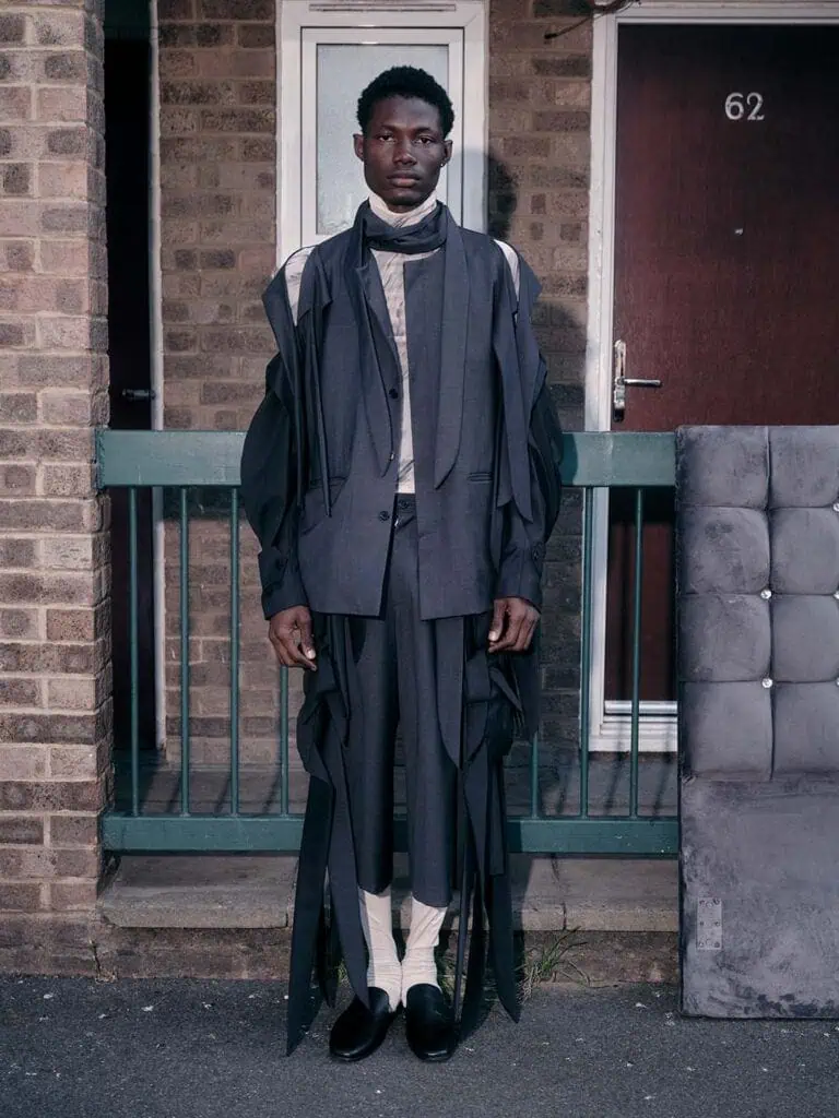 A man stands in front of a brick building and green railing, wearing avant-garde clothes designed by Yonzu—a layered gray suit with draping details, light shirt, white socks, and black shoes. A cushioned gray panel leans nearby. Door number 62 is visible.