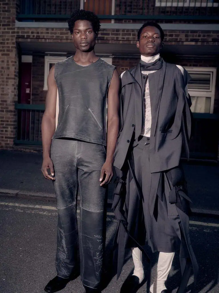 Two men stand side by side on a street, dressed in clothes designed by Yonzu—one in a sleeveless, distressed dark outfit, the other in a layered avant-garde black suit with long fabric strips and a white shirt. A brick building is in the background.