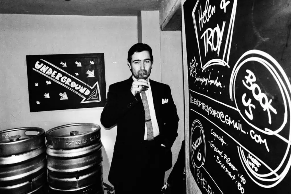 A man in a suit drinks from a glass in a bar-like setting, standing between kegs and a chalkboard with Book Us and contact info. A sign on the wall reads Underground, while "Tickety Boo" adds charm to the scene.