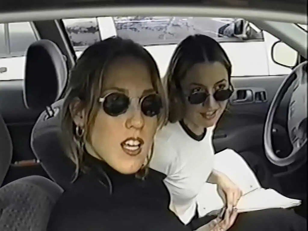 Two women wearing sunglasses sit in the front seats of a car, reminiscent of a scene from Dirty Girls documentary—one in black at the wheel and one in white smiling with open papers in the passenger seat.