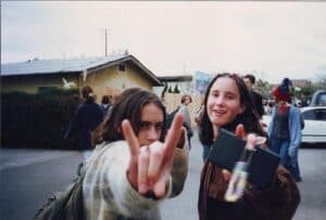 Two young people outdoors, one making a rock and roll hand gesture close to the camera while both smile, echoing the rebellious spirit of Dirty Girls documentary. Several people and cars are visible in the background on a cloudy day.