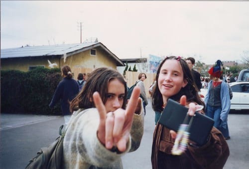 Two young people outdoors, one making a rock and roll hand gesture close to the camera while both smile, echoing the rebellious spirit of Dirty Girls documentary. Several people and cars are visible in the background on a cloudy day.