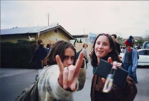 Two young people outdoors, one making a rock and roll hand gesture close to the camera while both smile, echoing the rebellious spirit of Dirty Girls documentary. Several people and cars are visible in the background on a cloudy day.