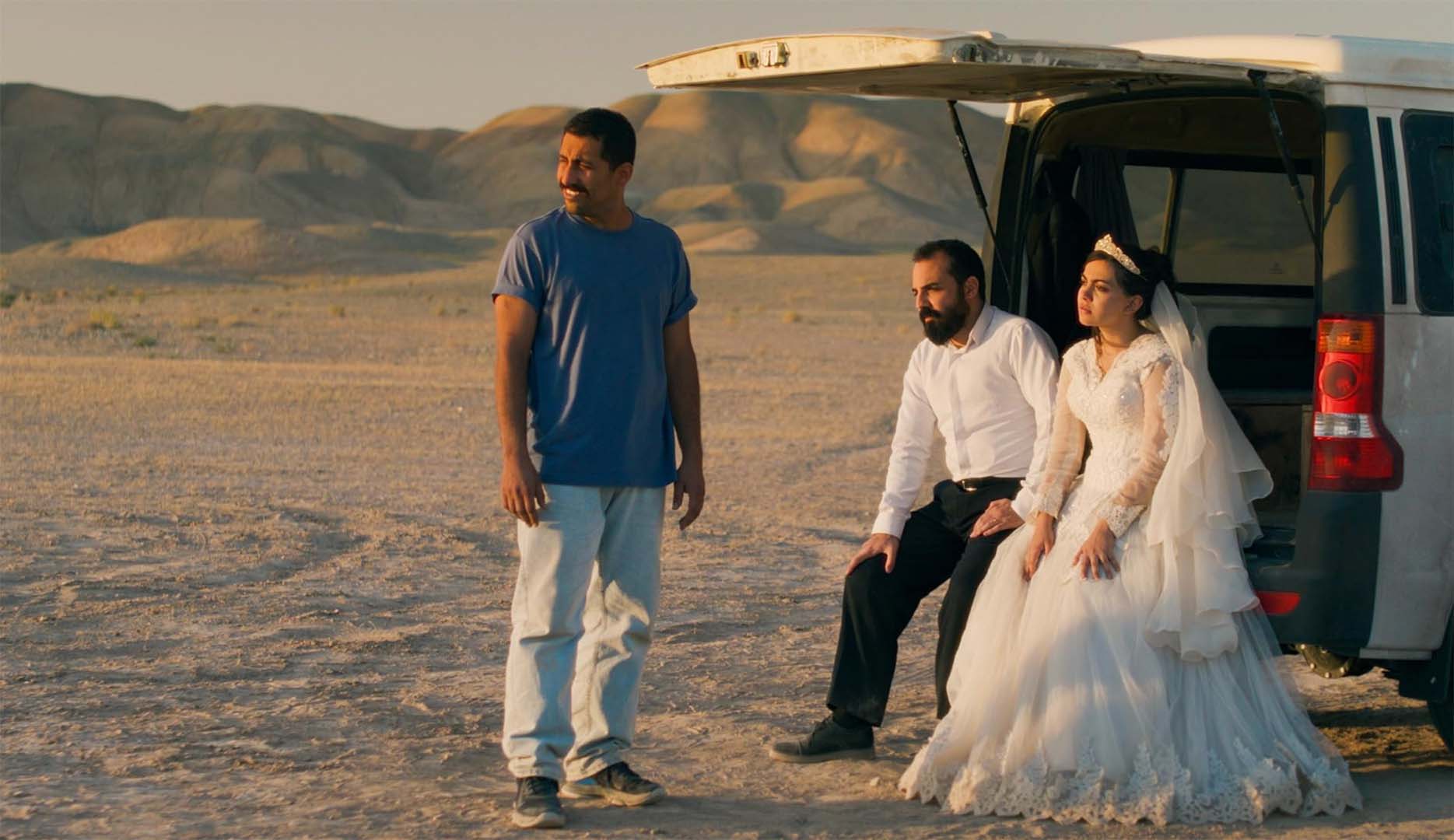 A bride and groom in wedding attire sit at the back of a white van in a desert, while a man in casual clothes stands nearby. Distant hills are visible under a clear sky—stills from movie ‘It Was Just an Accident’ by Jafar Panahi.