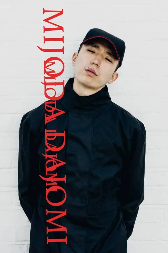 A young person wearing a black jacket and black cap designed by MIJODA DAJOMI stands against a white brick wall. Red vertical text reading MIJODA DAJOMI overlays the left side, hinting at 5 brands you should know for standout street style.