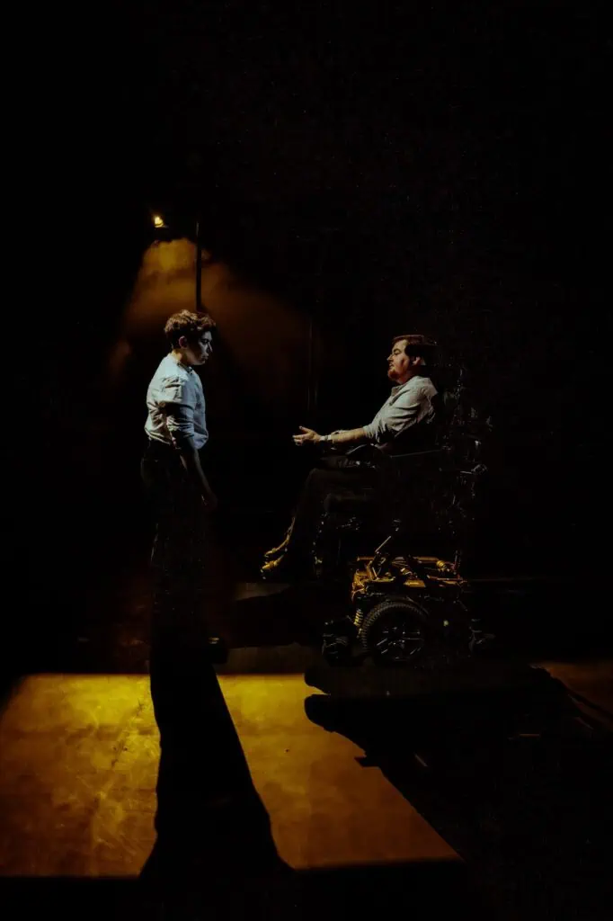Two men are dramatically lit on a dark stage at TRANSPOSE at the Barbican; one stands facing the other, who is sitting in a wheelchair. Shadows and warm light create a tense, theatrical atmosphere.
