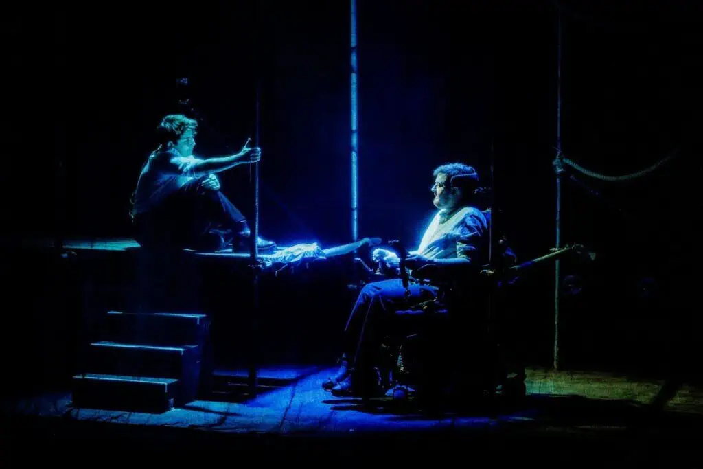 Two people perform on a dimly lit stage in TRANSPOSE at the Barbican. One sits on stairs holding a pole, while the other, in a wheelchair under a spotlight, faces them. The scene has a dramatic and moody atmosphere.