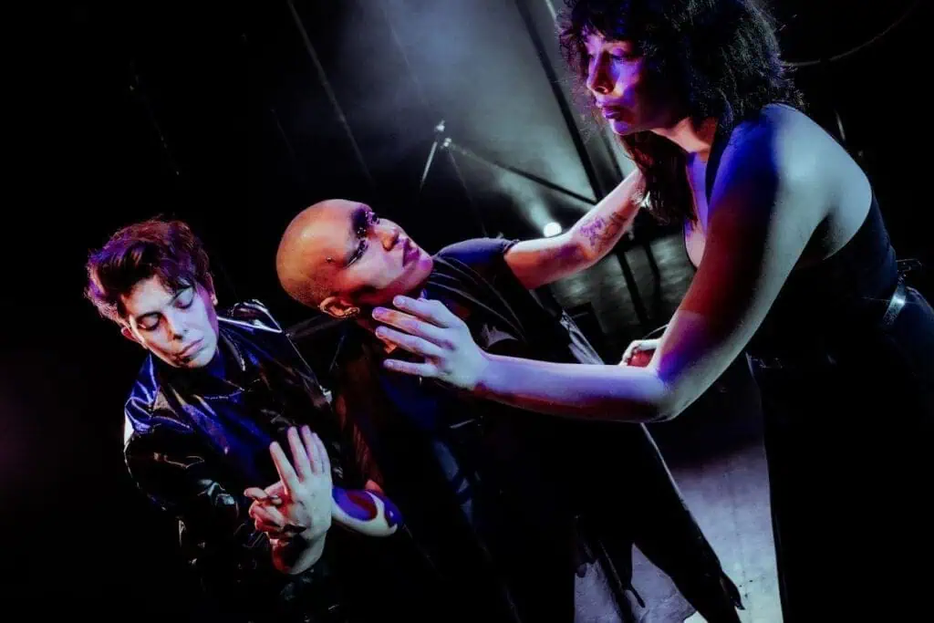 Three people on stage in dramatic lighting perform an intense scene at TRANSPOSE at the Barbican. Two reach out toward a third with long dark hair, all expressing strong emotion through gesture and facial expressions.