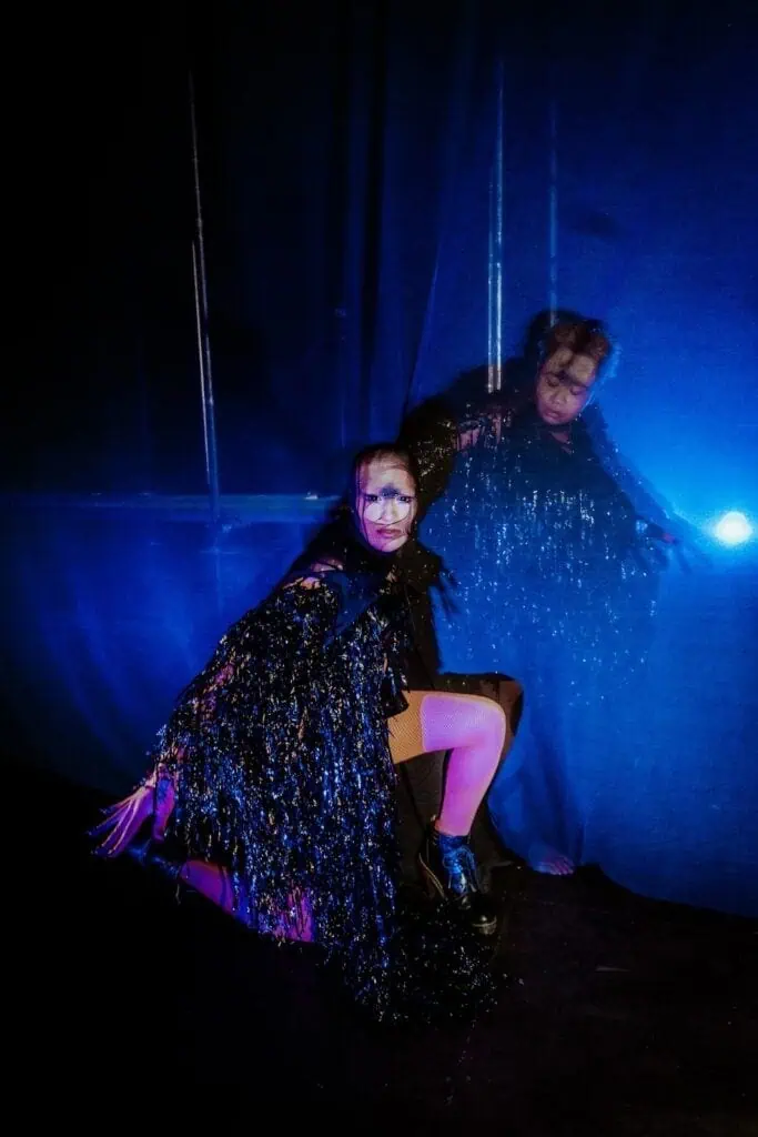 A person in dramatic makeup and a glittery, black fringed costume crouches against a dark blue lit background at TRANSPOSE at the Barbican, their reflection shimmering on a translucent surface. The scene is surreal and theatrical.
