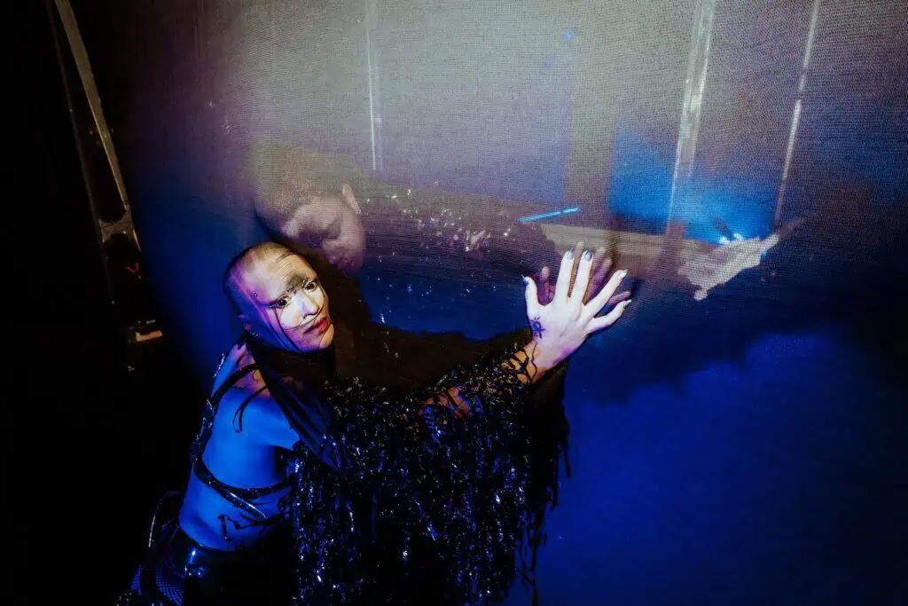 A person in dramatic black costume and makeup leans against a reflective surface at TRANSPOSE at the Barbican, pressing their hand to it. Their intense expression is echoed in their reflection within the blue-lit background.