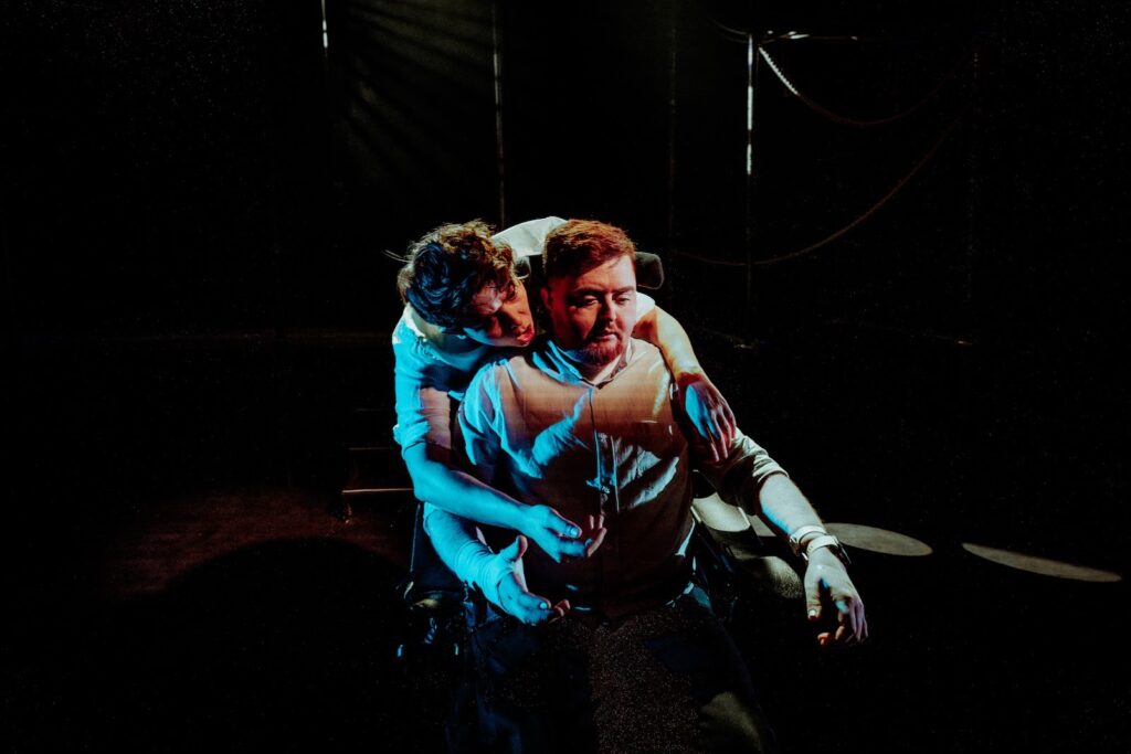 Two people sit in dramatic lighting at Transpose at the Barbican; one is seated while another leans over their shoulder, embracing them from behind. The dimly lit scene, with deep shadows, creates a tense and emotional atmosphere.