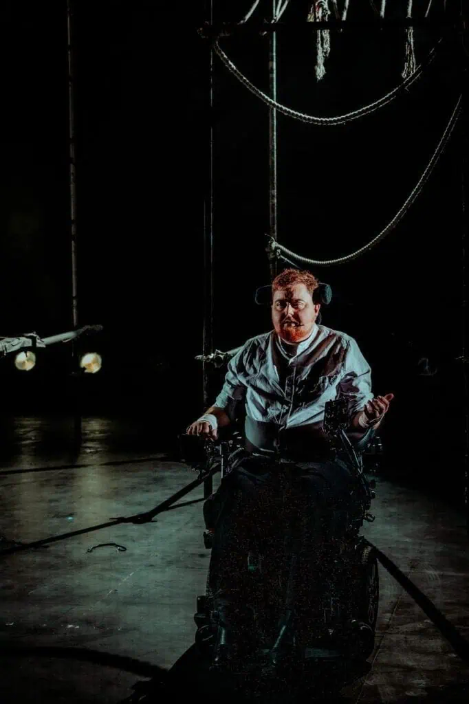 A person in a wheelchair sits on a dimly lit stage at TRANSPOSE at the Barbican, illuminated by dramatic lighting. Rope structures and spotlights in the background create a theatrical and intense atmosphere.