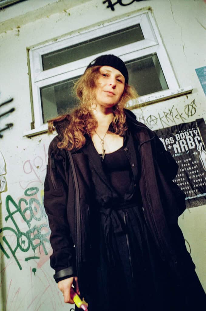 Maria Alyokhina with long curly hair, wearing a black beanie and dark layered clothing, stands in front of a graffiti-covered wall and a poster beneath a window, channeling Pussy Riot energy while looking confidently at the camera.
