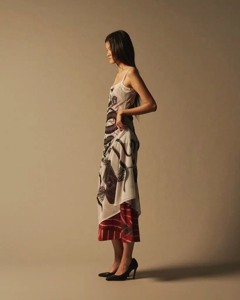 A woman stands in profile against a neutral background, wearing a sheer, patterned dress designed by Acceptance letter over a red skirt and black heels. Her long hair is down and her hands rest on her hips.