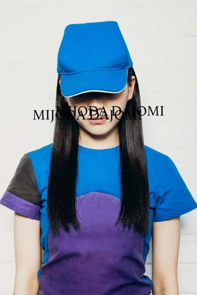 A person with long dark hair, wearing a blue cap designed by MIJODA DAJOMI and a blue and purple shirt, stands against a light-colored wall. Black text reading MIJSHADA JO MOMI is printed across the image, hinting at 5 brands you should know. The face is mostly obscured by the cap.