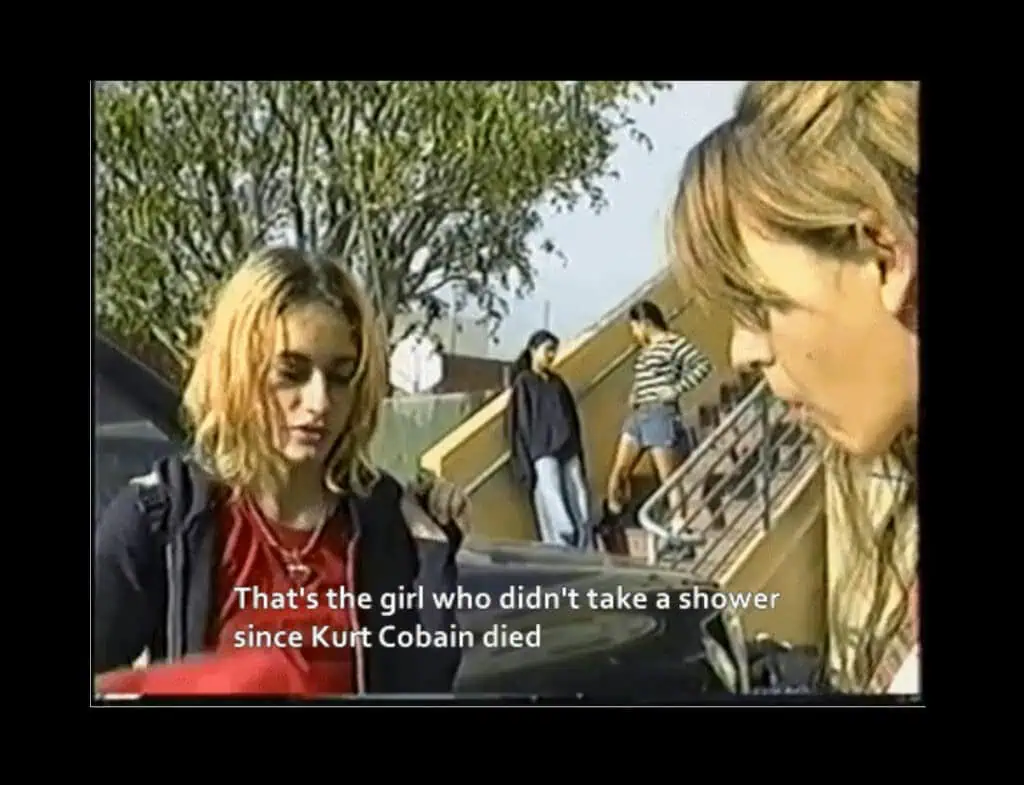 Two young women stand and talk outdoors near a staircase and tree. Caption reads, That’s the girl who didn’t take a shower since Kurt Cobain died. This scene is reminiscent of the Dirty Girls documentary, with two more people in the background.