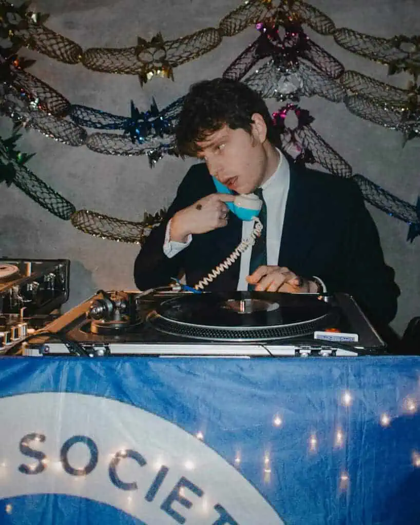 A man in a suit stands at a DJ booth, holding a blue rotary phone to his ear while adjusting a vinyl record. Colorful decorations and string lights create a Tickety Boo vibe behind him, adding to the lively atmosphere.