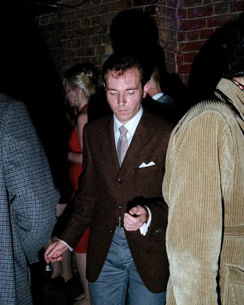 A man in a brown suit and patterned tie is dancing with his eyes closed at a dimly lit, Tickety Boo event. Other people are visible around him, including a woman in a red dress, all standing against a brick wall.