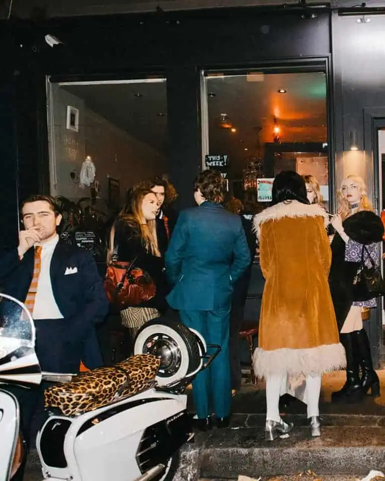 A stylish group of people gathers outside a dimly lit café at night; one Tickety Boo man in a suit stands near a white scooter with a leopard print seat, while others chat by the entrance, dressed in fashionable retro clothing.